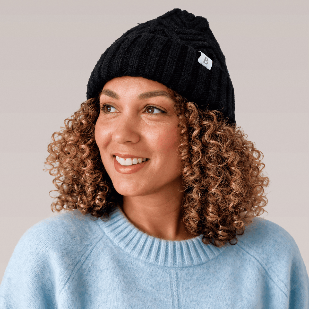 A smiling woman with curly brown hair wearing the Bouclème black Silk Lined Beanie with a light blue sweater, photographed against a plain light grey background.