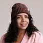A close-up portrait of a woman with curly hair wearing a hot chocolate Bouclème Silk Lined Beanie set against a solid light grey background.