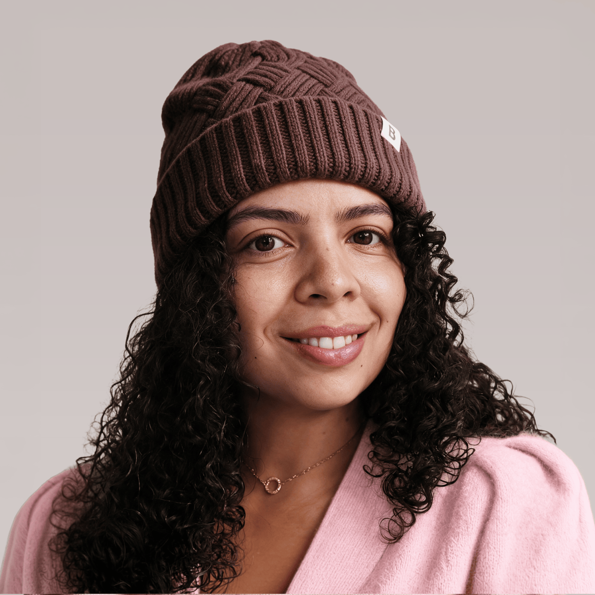 A close-up portrait of a woman with curly hair wearing a hot chocolate Bouclème Silk Lined Beanie set against a solid light grey background.