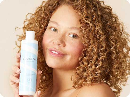 woman with strawberry-blonde hair holding a bottle of Bouclème's Hydrating Hair Cleanser