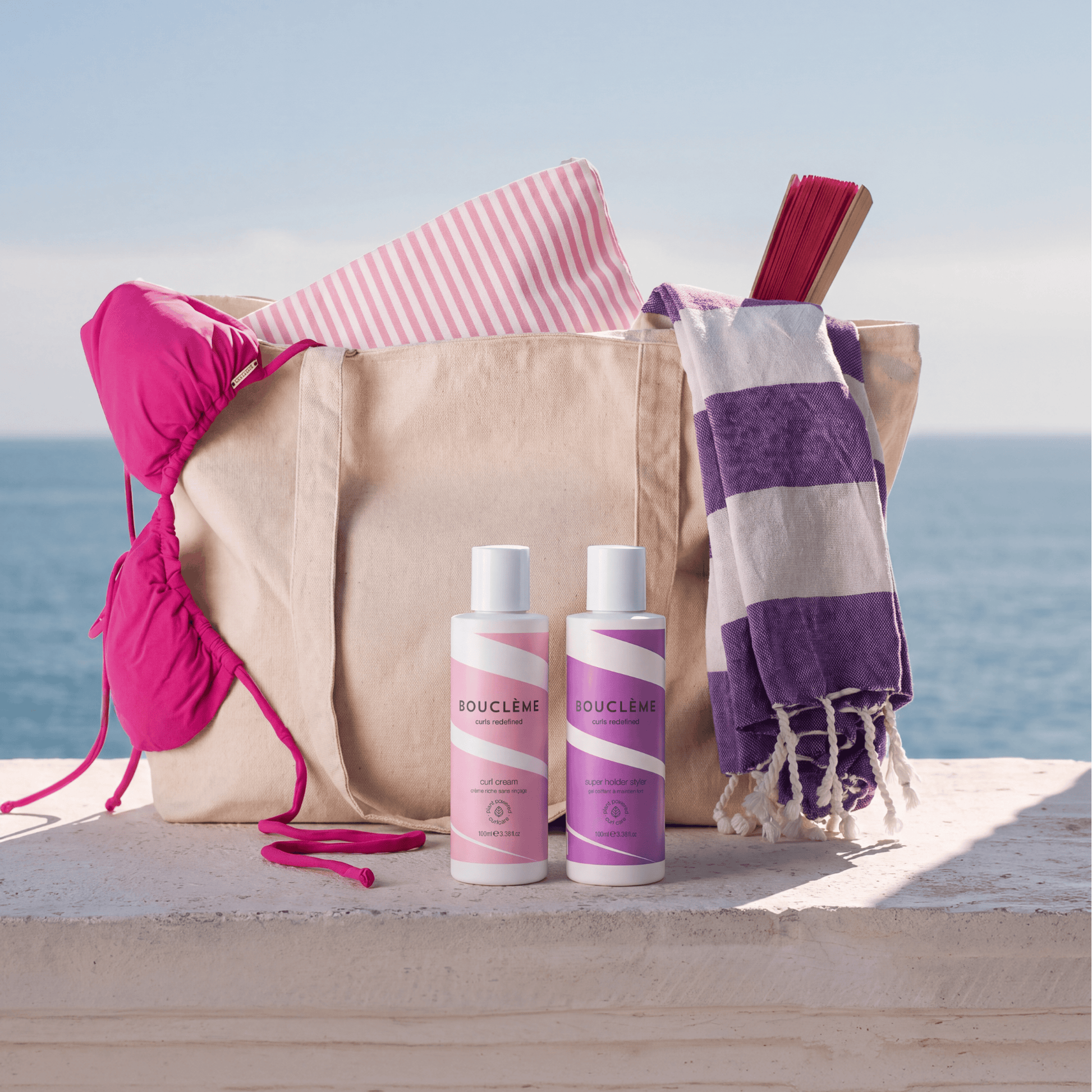 Bouclemi Hydration & Hold Duo displayed in front of a beige tote bag with beach accessories, including a pink bikini top, striped towel, and purple-and-white fringed towel, set against an ocean background.